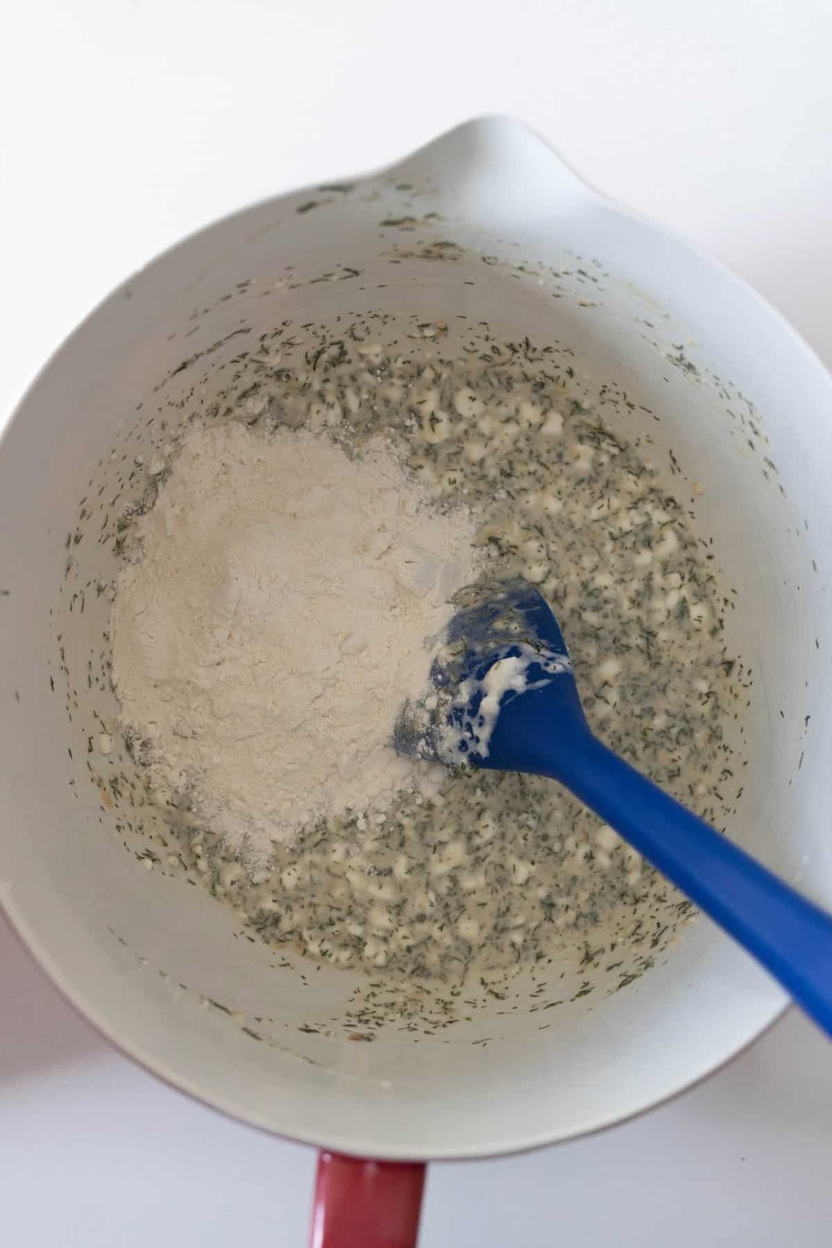 A mixing bowl with a blue spatula contains a mixture of chopped herbs and liquid, with flour partially added on top—perfect for starting your Dilly Bread Recipe.
