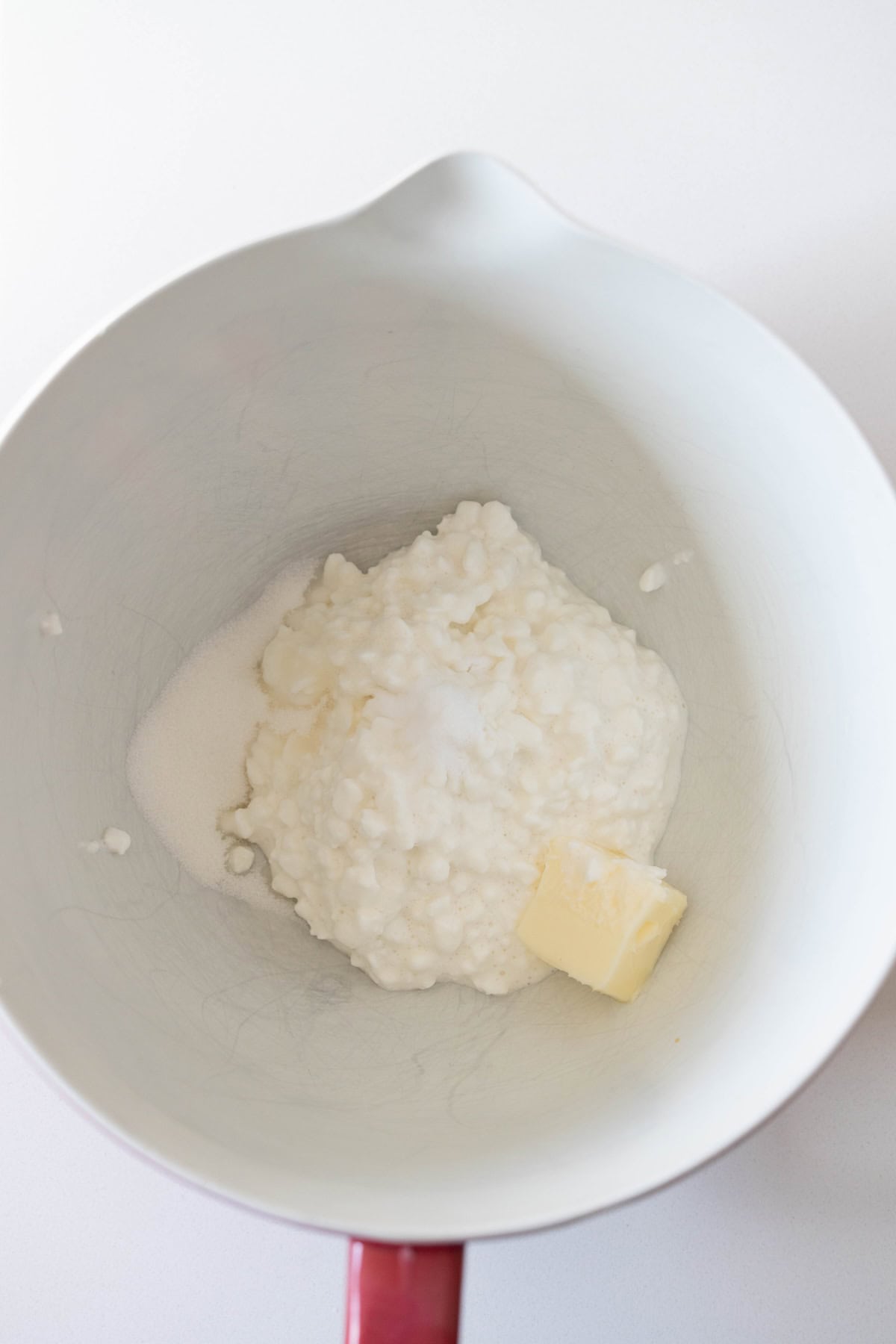 A mixing bowl containing cottage cheese, a pat of butter, and a small pile of granulated sugar on a white surface—perfect beginnings for a classic dill bread recipe.