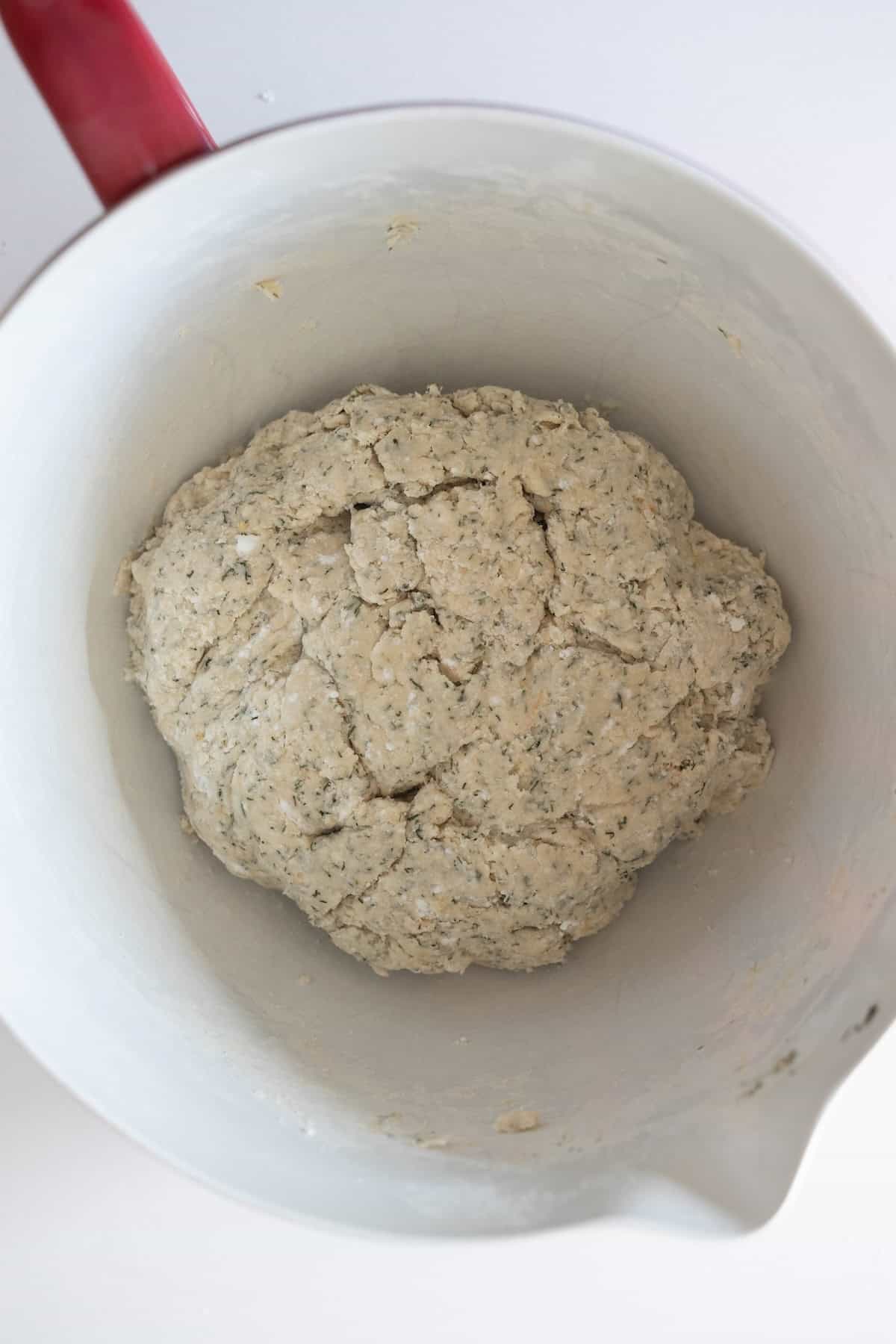 A ball of herbed dough for dilly bread sits in a white mixing bowl with a red handle, ready for further preparation.