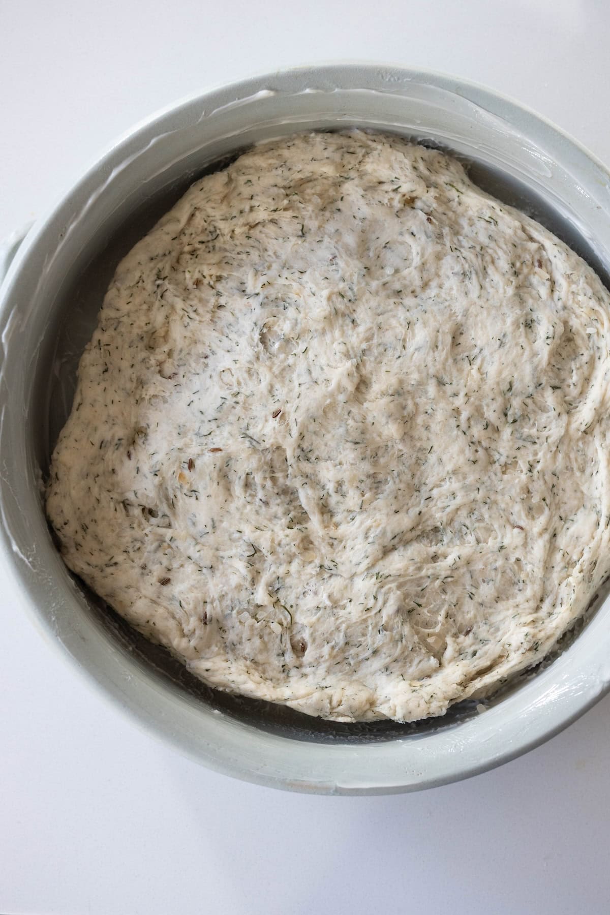 A bowl filled with risen, herb-speckled dough for a classic Dilly Bread Recipe sits on a white surface.