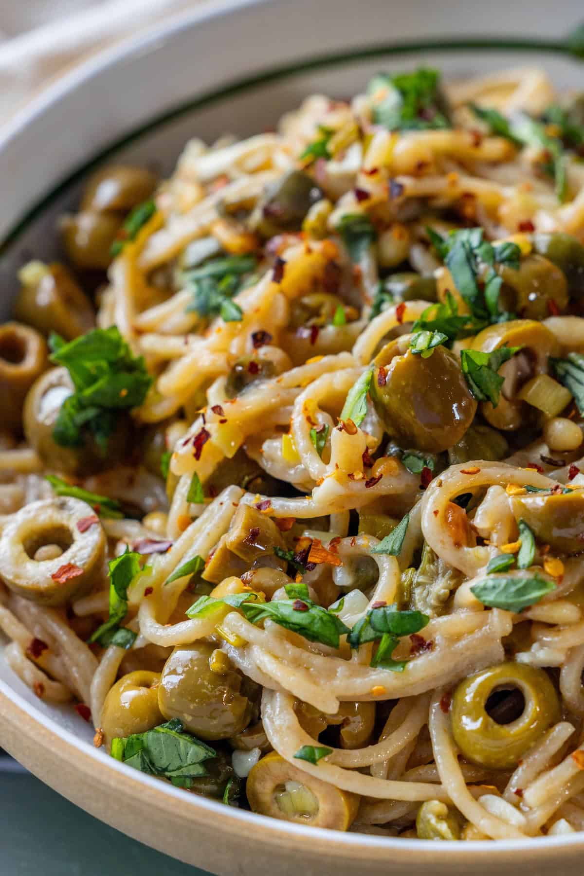 A close-up of spaghetti puttanesca mixed with sliced green olives, chopped parsley, red pepper flakes, and a light sauce in a bowl.