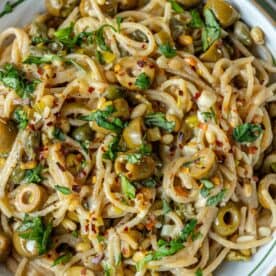 Close-up of spaghetti puttanesca with green olives, fresh herbs, red pepper flakes, and a creamy sauce, served in a white bowl.