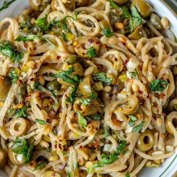 Close-up of spaghetti puttanesca with green olives, fresh herbs, red pepper flakes, and a creamy sauce, served in a white bowl.