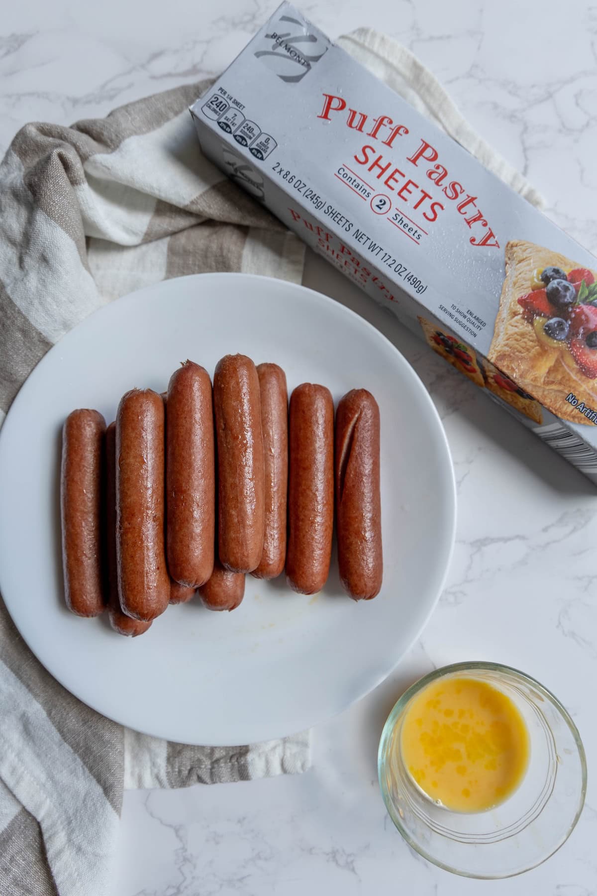 A white plate with several mummy hot dogs, a bowl of beaten egg, and a box of puff pastry sheets on a striped towel.