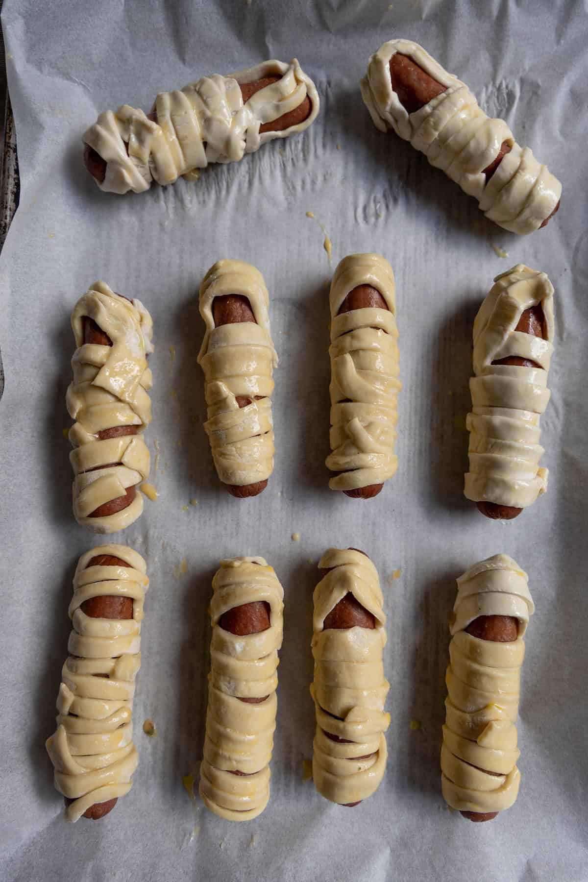 Nine mummy hot dogs, wrapped in strips of dough, are arranged on a parchment-lined baking sheet, ready to be baked.