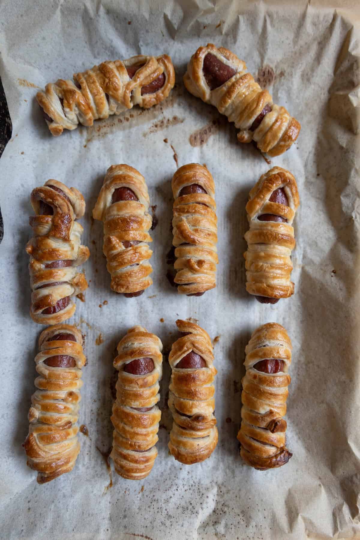 Eight sausage rolls wrapped in puff pastry, arranged on a parchment-lined baking tray. Reminiscent of mummy hot dogs, the pastry is golden brown and slightly flaky.