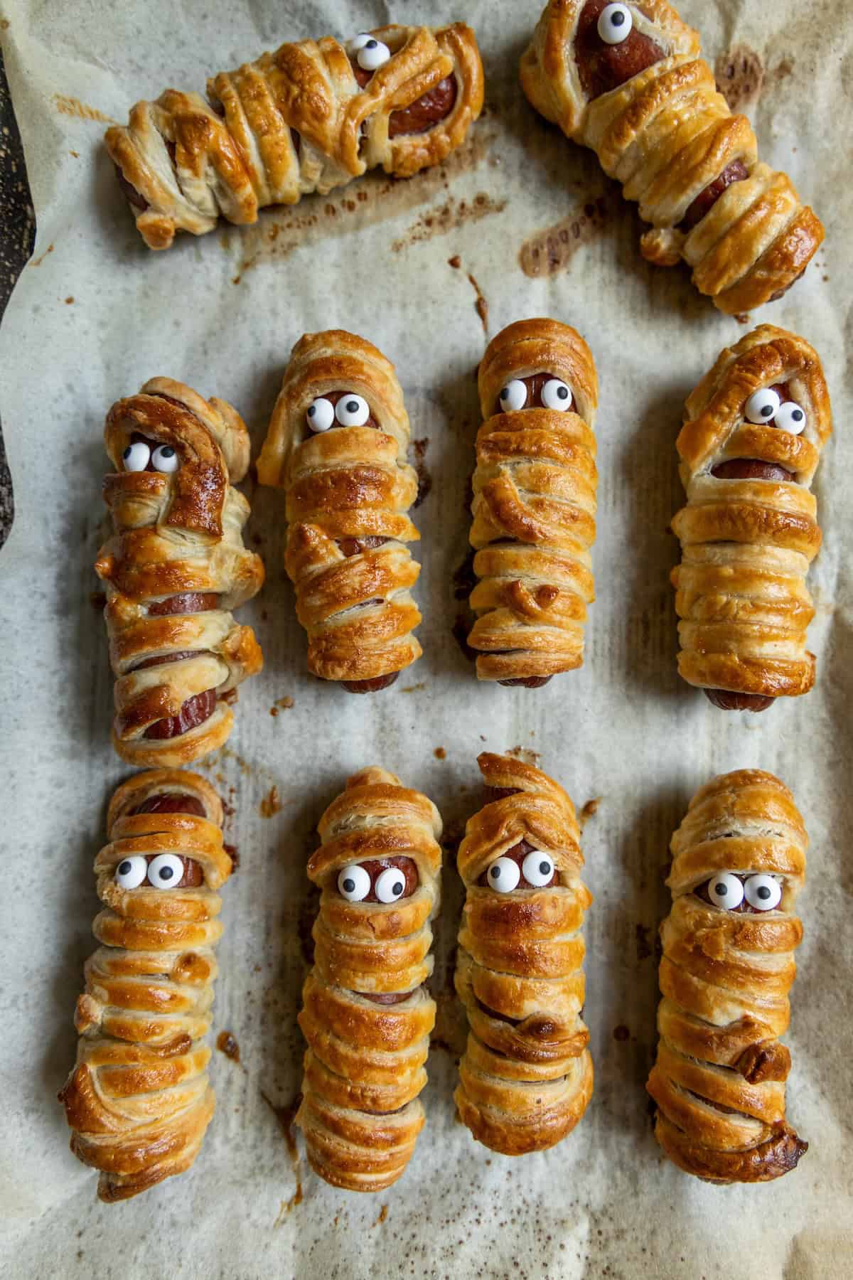 Rows of mummy hot dogs wrapped in pastry and topped with candy eyes are arranged on a parchment-lined baking tray, creating a spooky, festive treat.