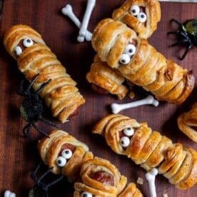 Mummy hot dogs wrapped in pastry to look like mummies with candy eyes, surrounded by fake spiders and bone decorations on a wooden surface.