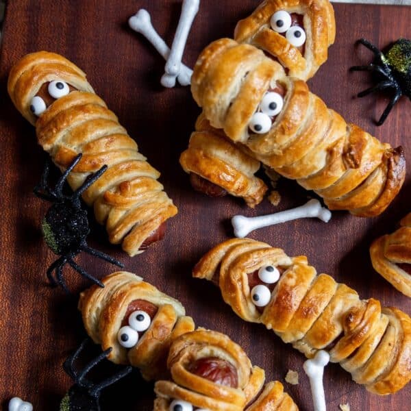 Mummy hot dogs wrapped in pastry to look like mummies with candy eyes, surrounded by fake spiders and bone decorations on a wooden surface.