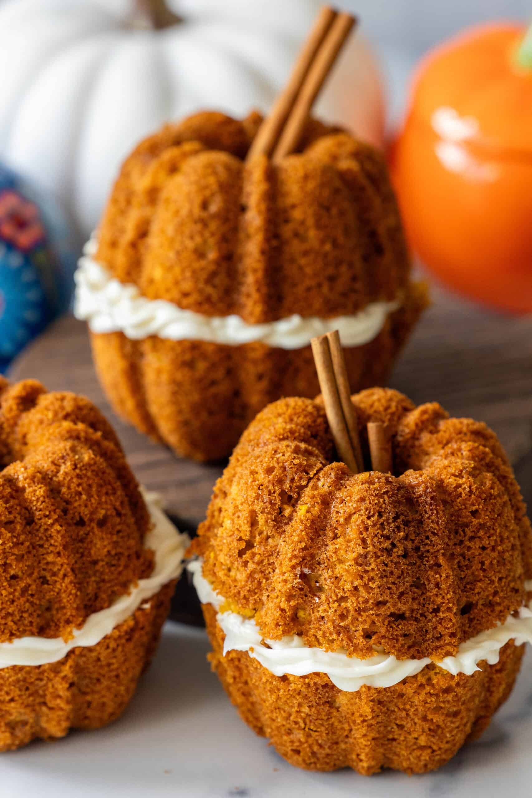 Three pumpkin-shaped cakes with cream filling in the center, each topped with cinnamon sticks, displayed on a table with pumpkins in the background.