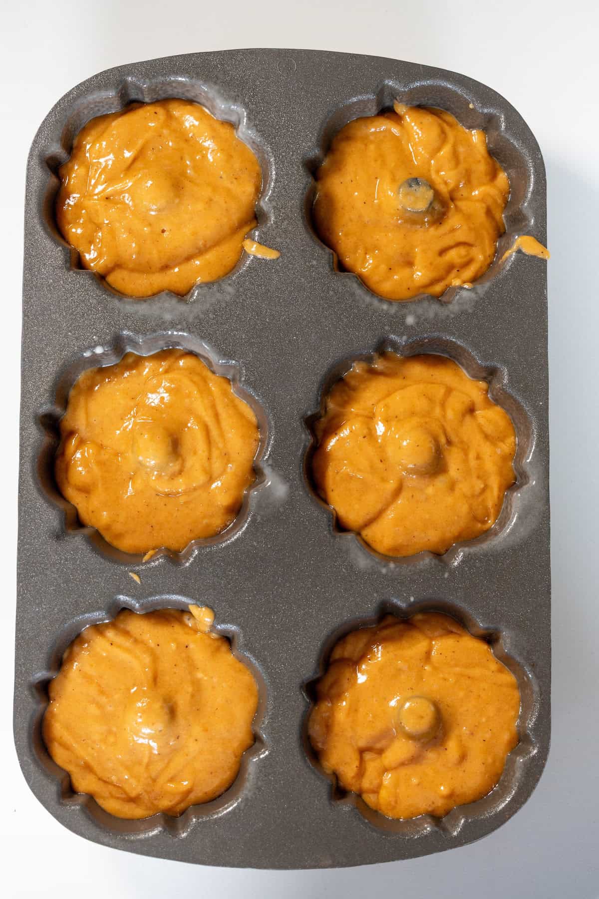 A six-cavity donut baking pan filled with unbaked orange batter, ready to be placed in the oven.