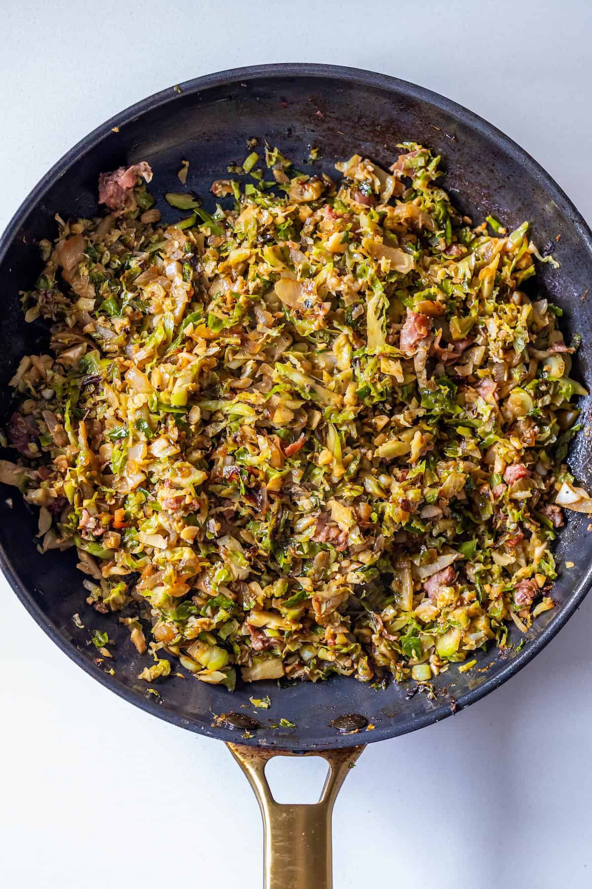 A skillet filled with savory Brussels Sprouts Hash, featuring cooked, shredded Brussels sprouts mixed with bacon and herbs, sits on a light background.