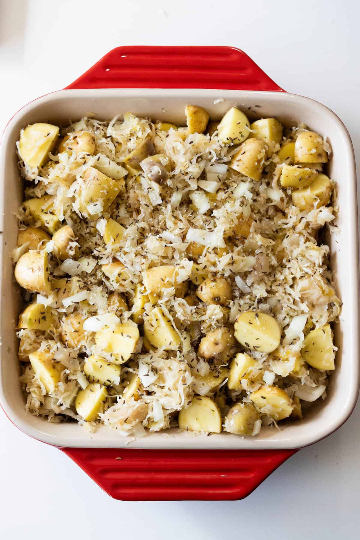A square red baking dish filled with roasted potatoes and sauerkraut, seasoned with herbs, sits on a white surface.