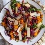A plate of winter salad with kale, roasted butternut squash, red onions, purple cabbage, pomegranate seeds, walnuts, and shaved cheese.