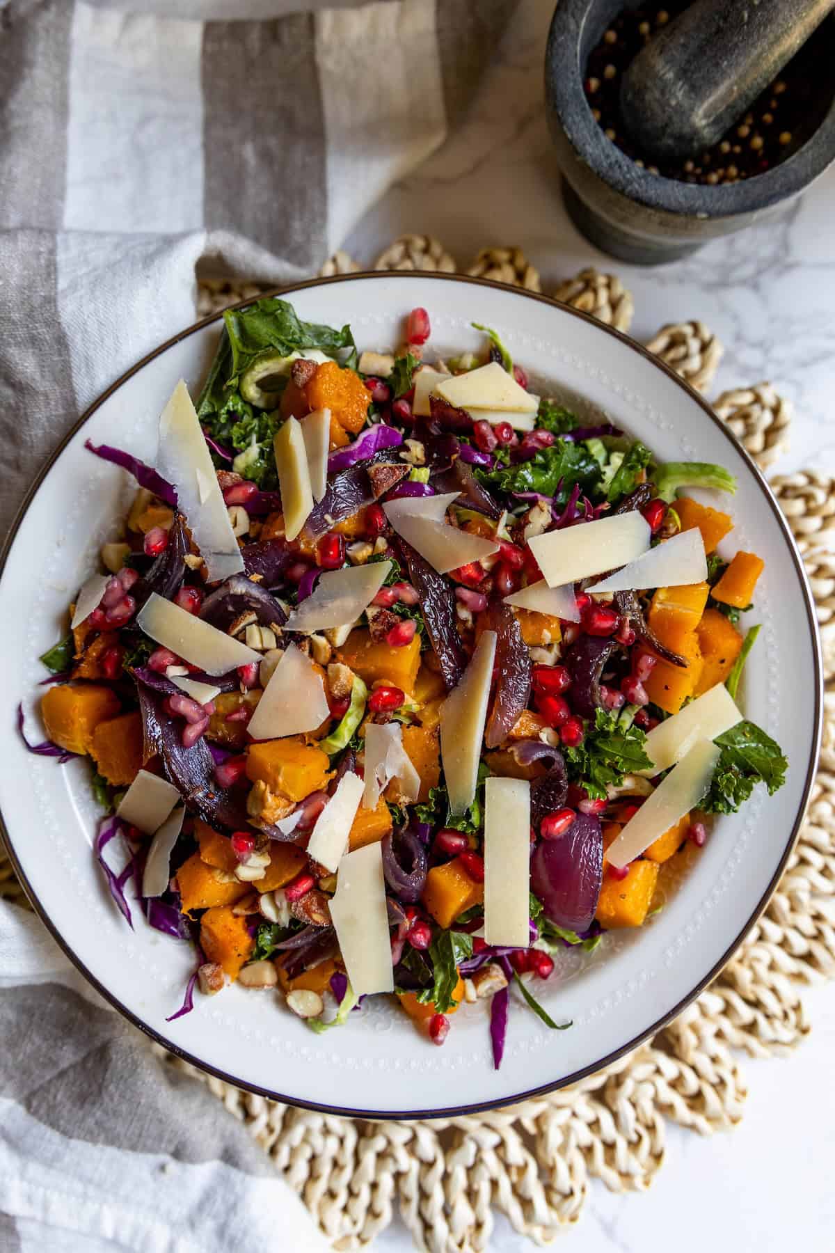 A vibrant winter salad with mixed greens, roasted butternut squash, red onion, pomegranate seeds, pumpkin seeds, and shaved cheese on a white plate. A mortar with peppercorns sits nearby.