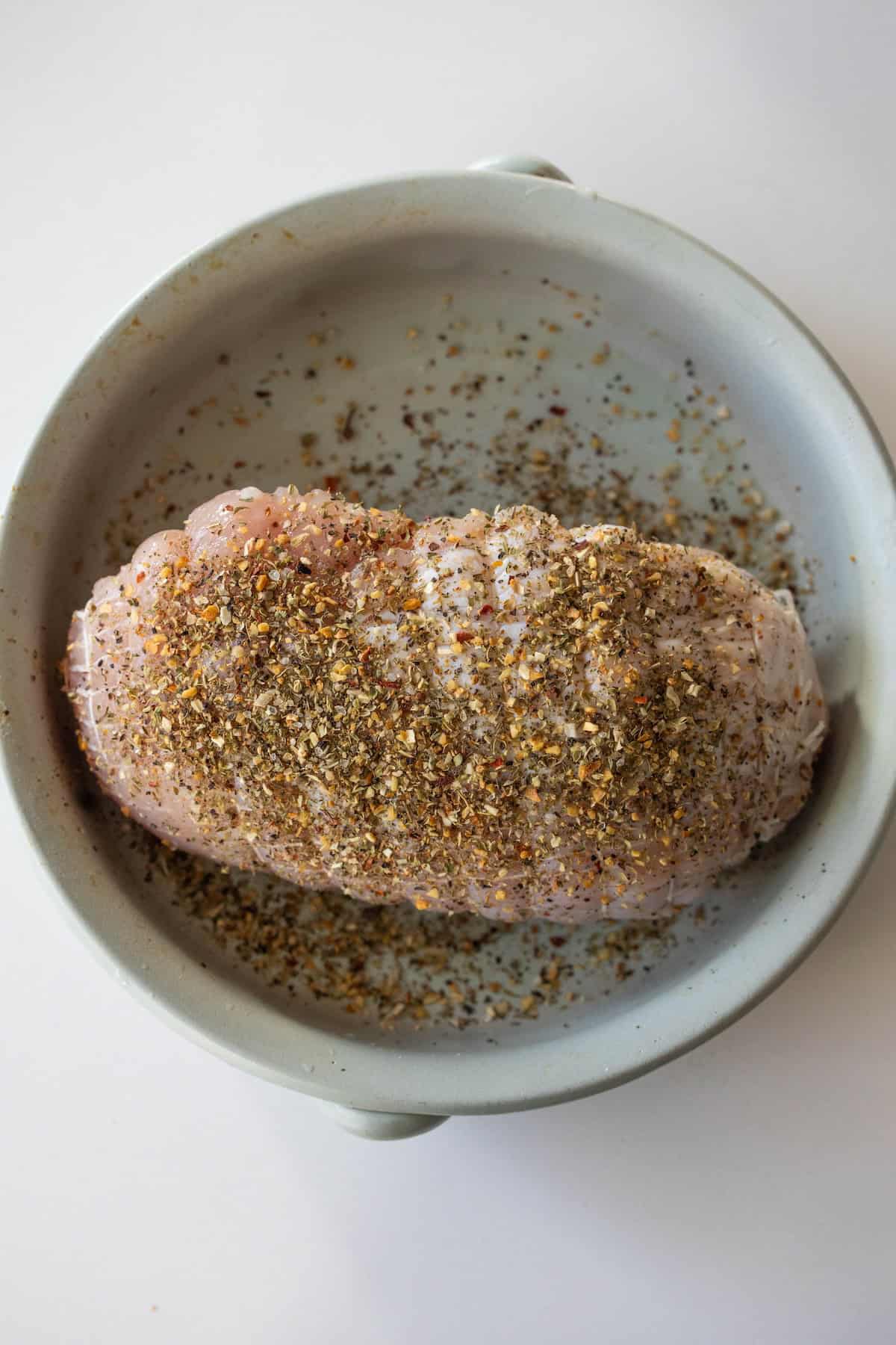 A raw, netted turkey breast coated with a mixture of spices sits in a round, light-colored baking dish on a white surface, ready to be cooked in the air fryer for a flavorful and juicy main course.