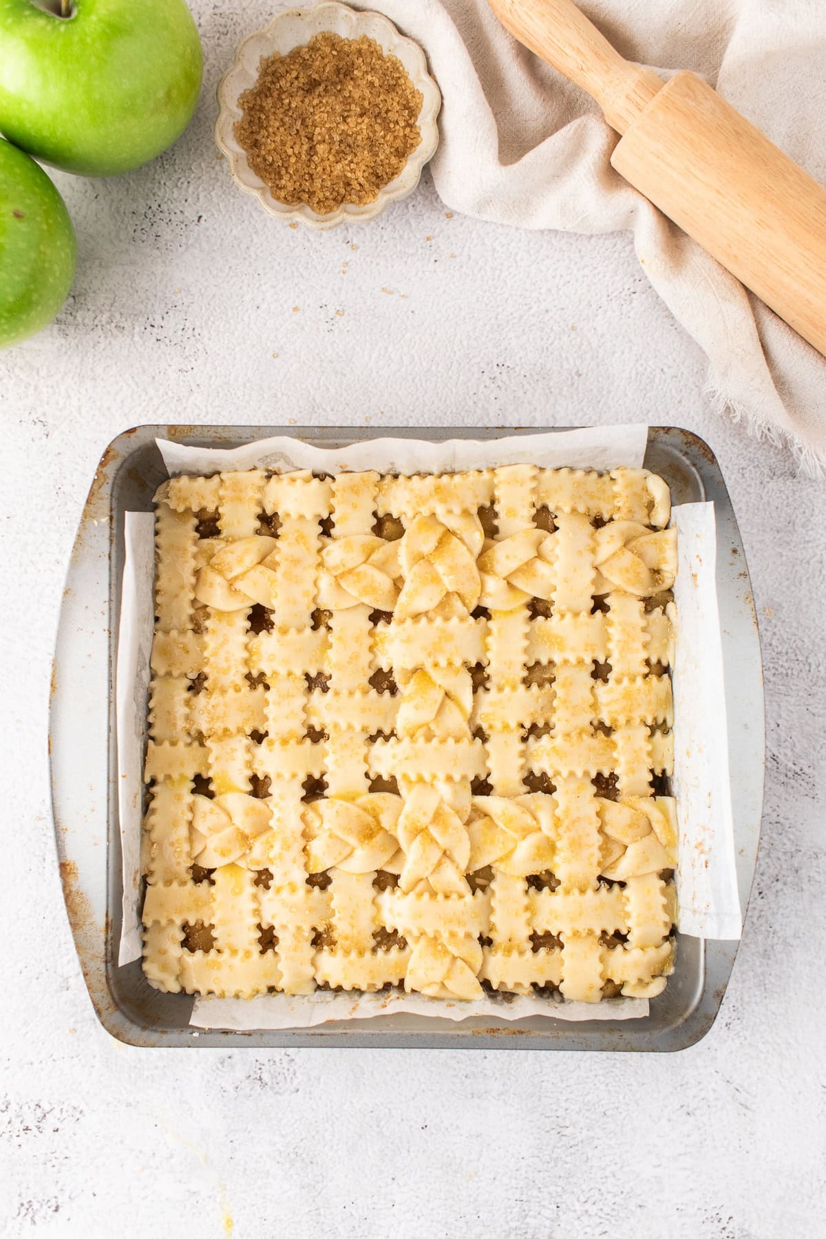 Square baking pan with unbaked Apple Pie Bars, decorated with braided and leaf-shaped dough. Surrounding items include green apples, brown sugar, a rolling pin, and a beige cloth—perfect for creating delicious dessert bars.