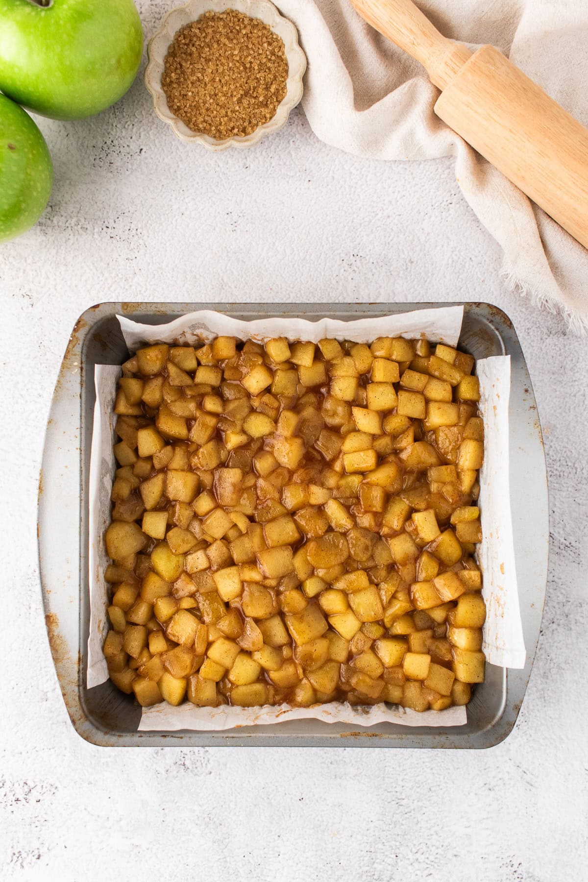 Baking pan filled with diced, cooked apples in a brown spiced mixture for Apple Pie Bars, lined with parchment paper, next to green apples, brown sugar, and a wooden rolling pin.