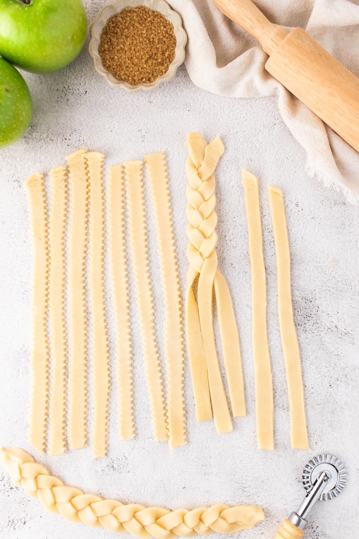 Unbaked pie crust strips, including a braided section, are arranged on a light surface with a rolling pin, green apples, brown sugar, and a pastry wheel nearby—perfect for making delicious Apple Pie Bars.