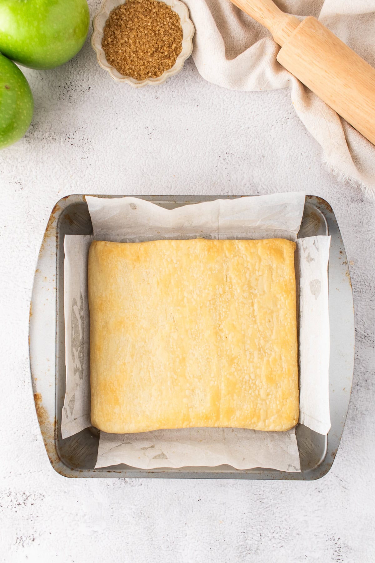 A square sheet of pastry dough lined in a parchment-lined baking pan, with a rolling pin, brown sugar, and green apples nearby—perfect for making delicious Apple Pie Bars.