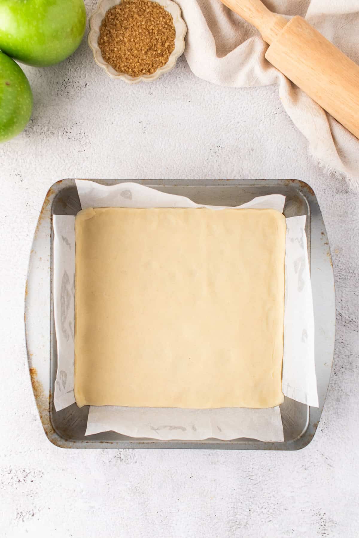 A square sheet of raw pastry dough is placed in a parchment-lined baking pan, ready to become delicious Apple Pie Bars. Nearby are green apples, a bowl of brown sugar, a rolling pin, and a beige cloth.