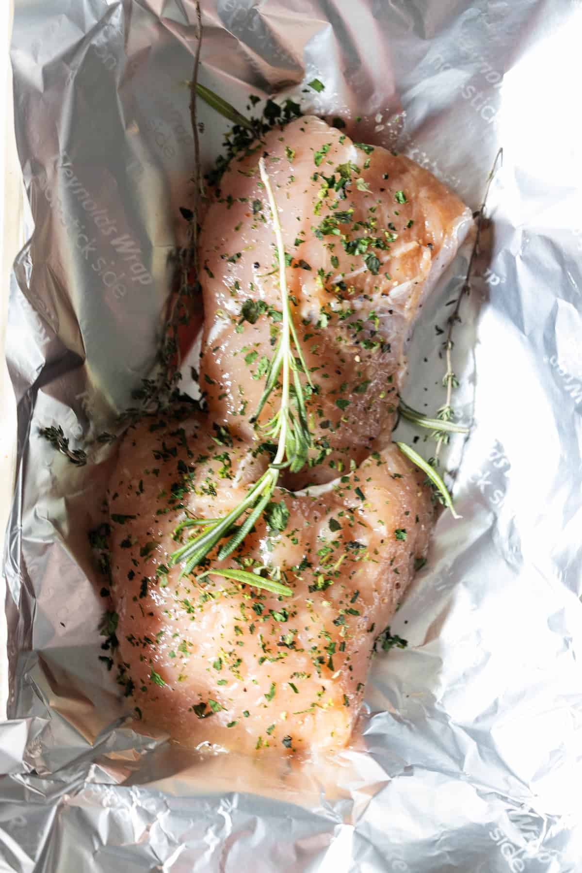 Raw chicken breasts seasoned with herbs and a sprig of rosemary, placed on a sheet of aluminum foil—just like preparing an easy turkey recipe or baking turkey tenderloin for a flavorful meal.