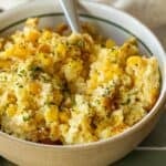 A bowl of scrambled eggs mixed with corn, inspired by classic Corn Pudding, garnished with chopped herbs, with a spoon resting inside the bowl.