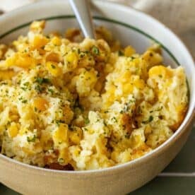 A bowl of scrambled eggs mixed with corn, inspired by classic Corn Pudding, garnished with chopped herbs, with a spoon resting inside the bowl.
