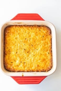 A baked casserole with a golden, cheesy top sits in a square red and white ceramic dish on a white background, showcasing this classic corn pudding recipe.
