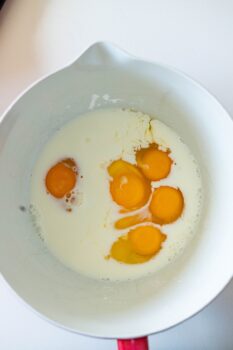A white mixing bowl contains six raw eggs and milk, ready to be whisked together as the base for a classic corn pudding recipe, all set on a clean white surface.