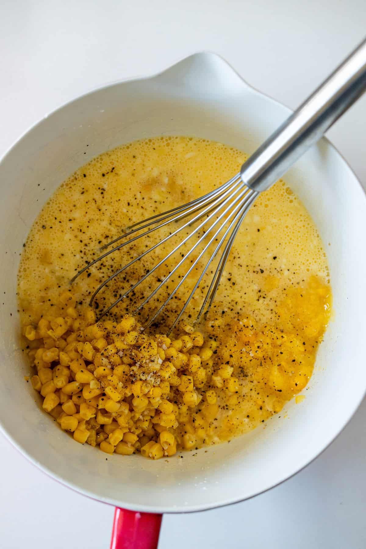 A whisk in a white mixing bowl with beaten eggs, corn kernels, and ground black pepper, ready to create an Easy Corn Pudding.