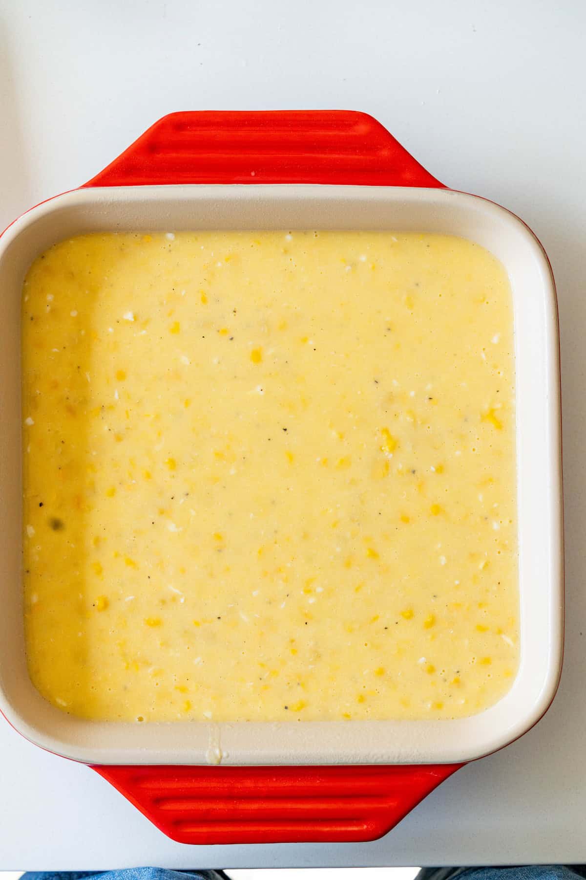 A red and white square baking dish filled with uncooked cornbread batter for an easy corn pudding sits on a white surface.