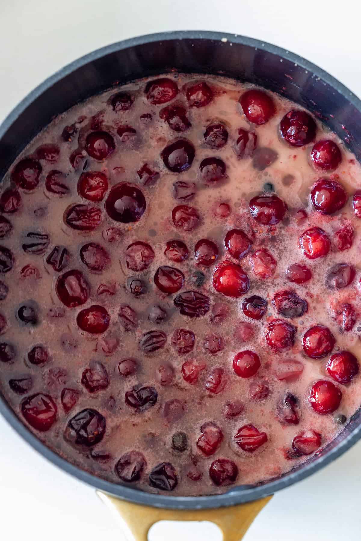 A pot filled with simmering cranberries in liquid, some berries whole and some burst, creates a foamy surface—perfect for glazing a Spiral Ham or adding a hint of Cranberry Brandy to your holiday feast.