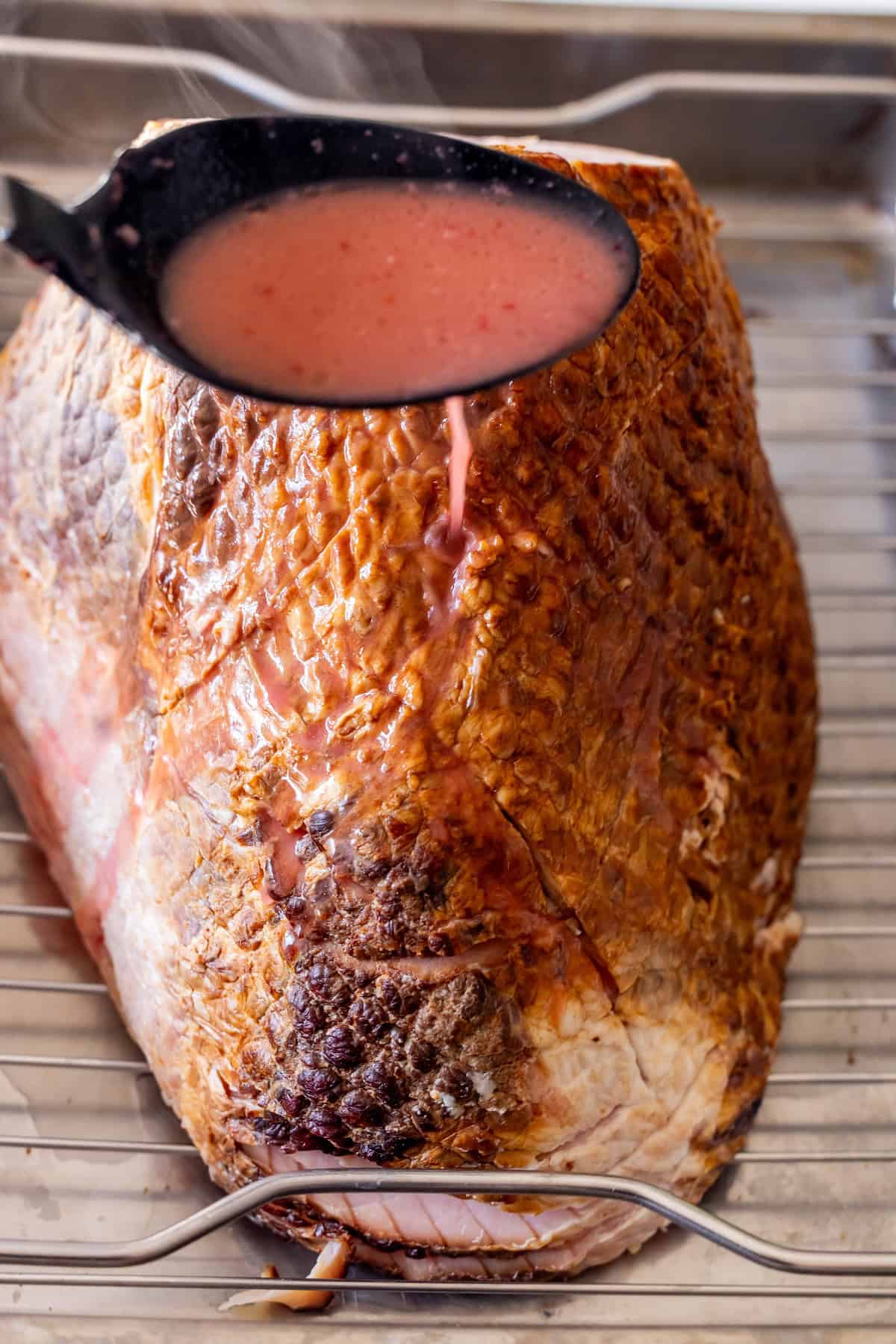A ladle pours pink Cranberry Brandy sauce over a large Glazed Spiral Ham resting on a wire rack in a roasting pan.