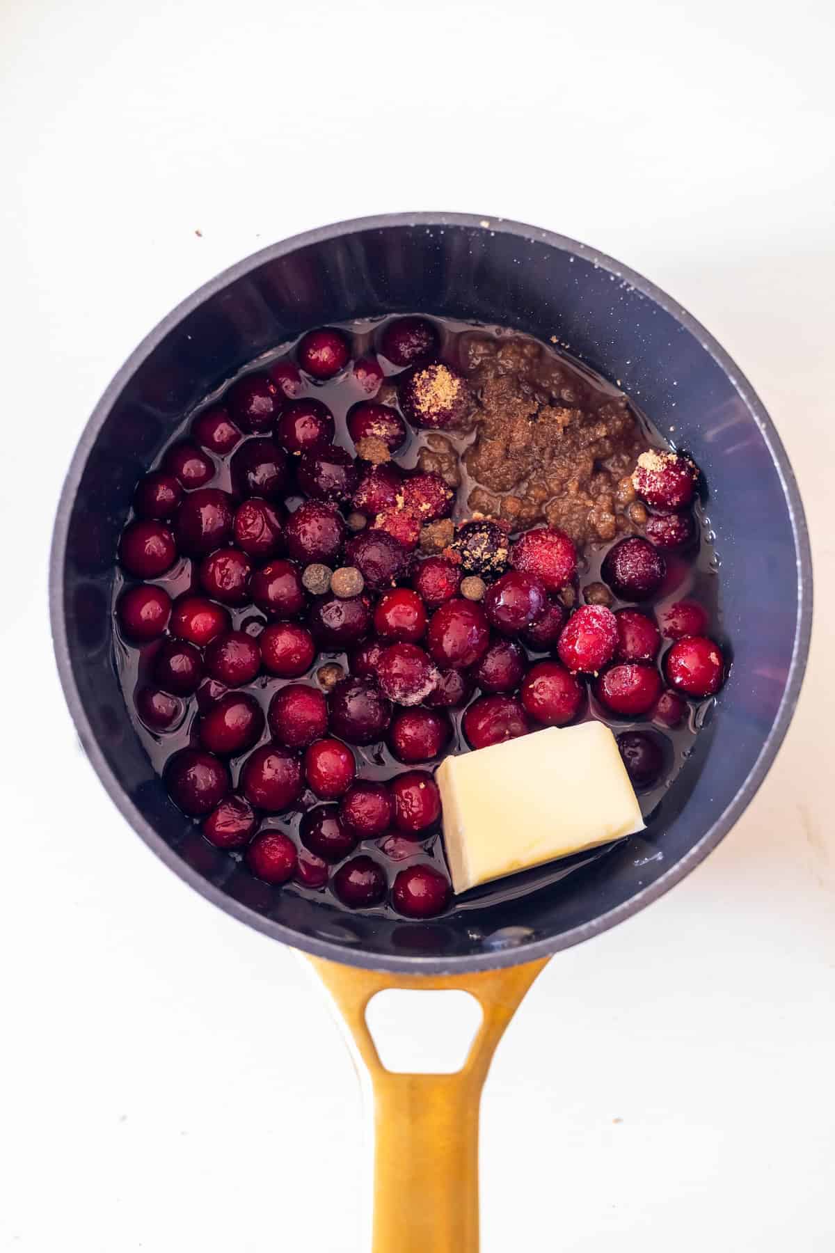 A saucepan containing whole cranberries, a pat of butter, brown sugar, and spices, ready to be cooked into a perfect topping for Glazed Ham.