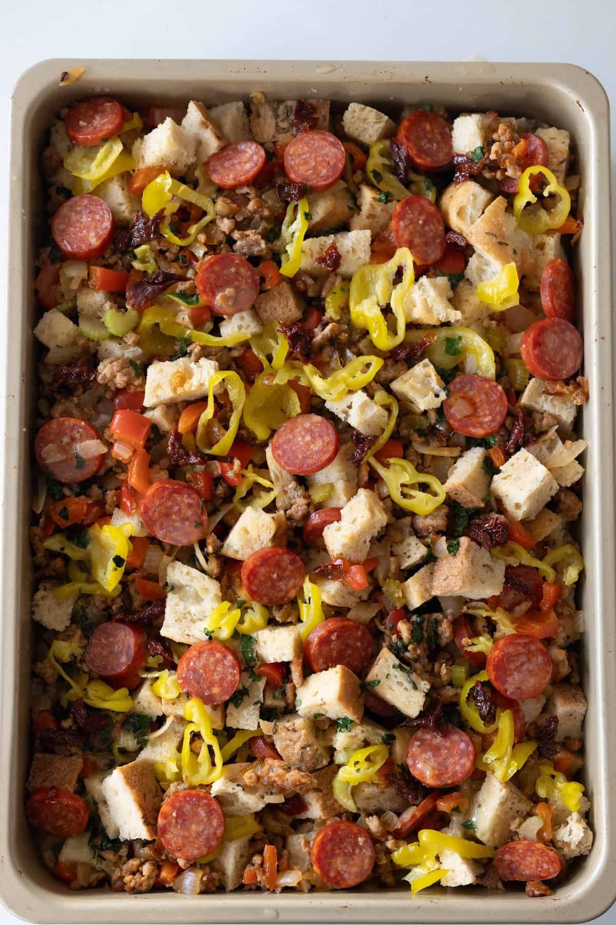 A baking dish filled with Pepperoni Stuffing—a layered mixture of cubed bread, pepperoni slices, banana peppers, ground meat, and chopped vegetables.