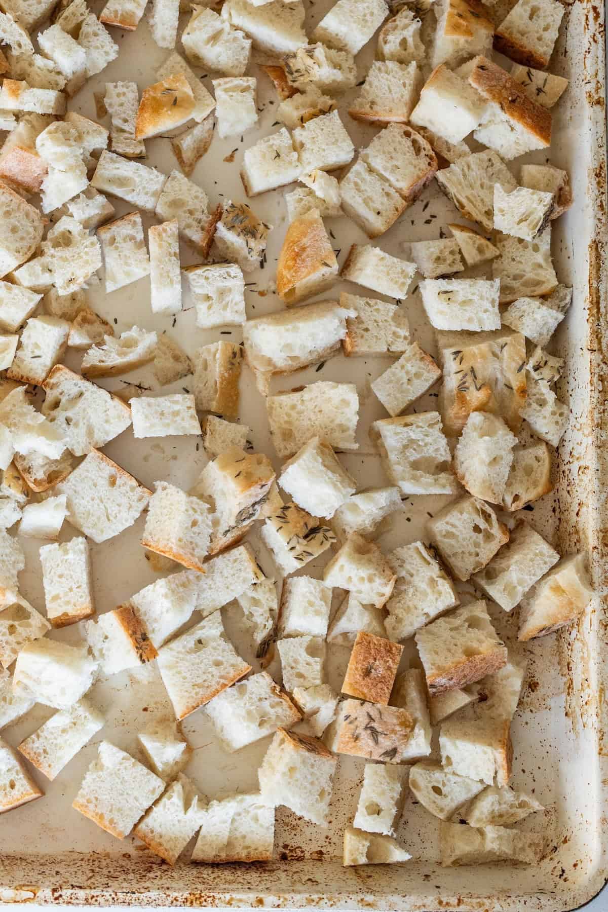 Cubed pieces of bread scattered on a baking sheet, prepared for toasting or drying, perfect as a base for savory stuffing or even a zesty pepperoni stuffing.