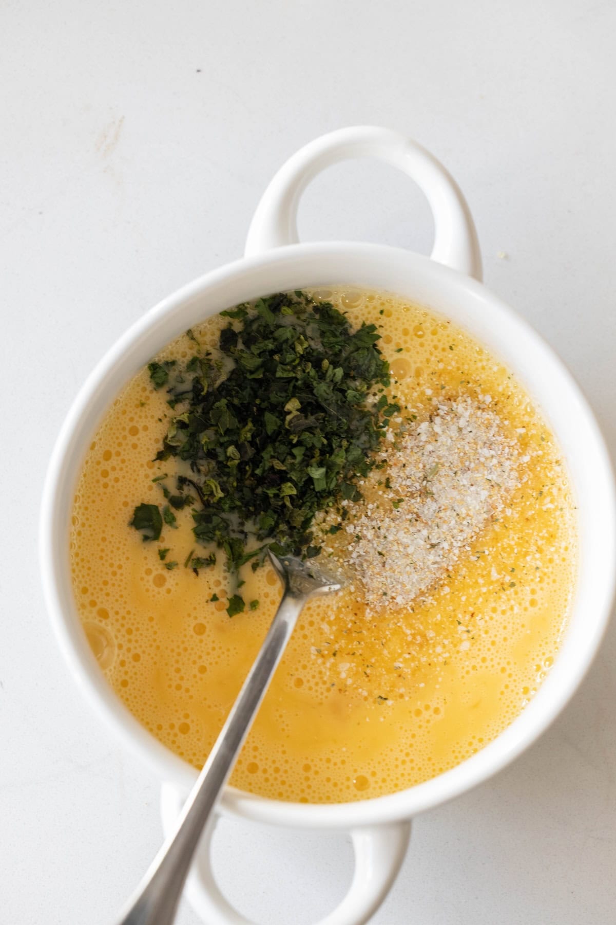 White bowl containing beaten eggs, chopped herbs, pepperoni stuffing, and seasoning, with a metal fork resting inside, on a white surface.