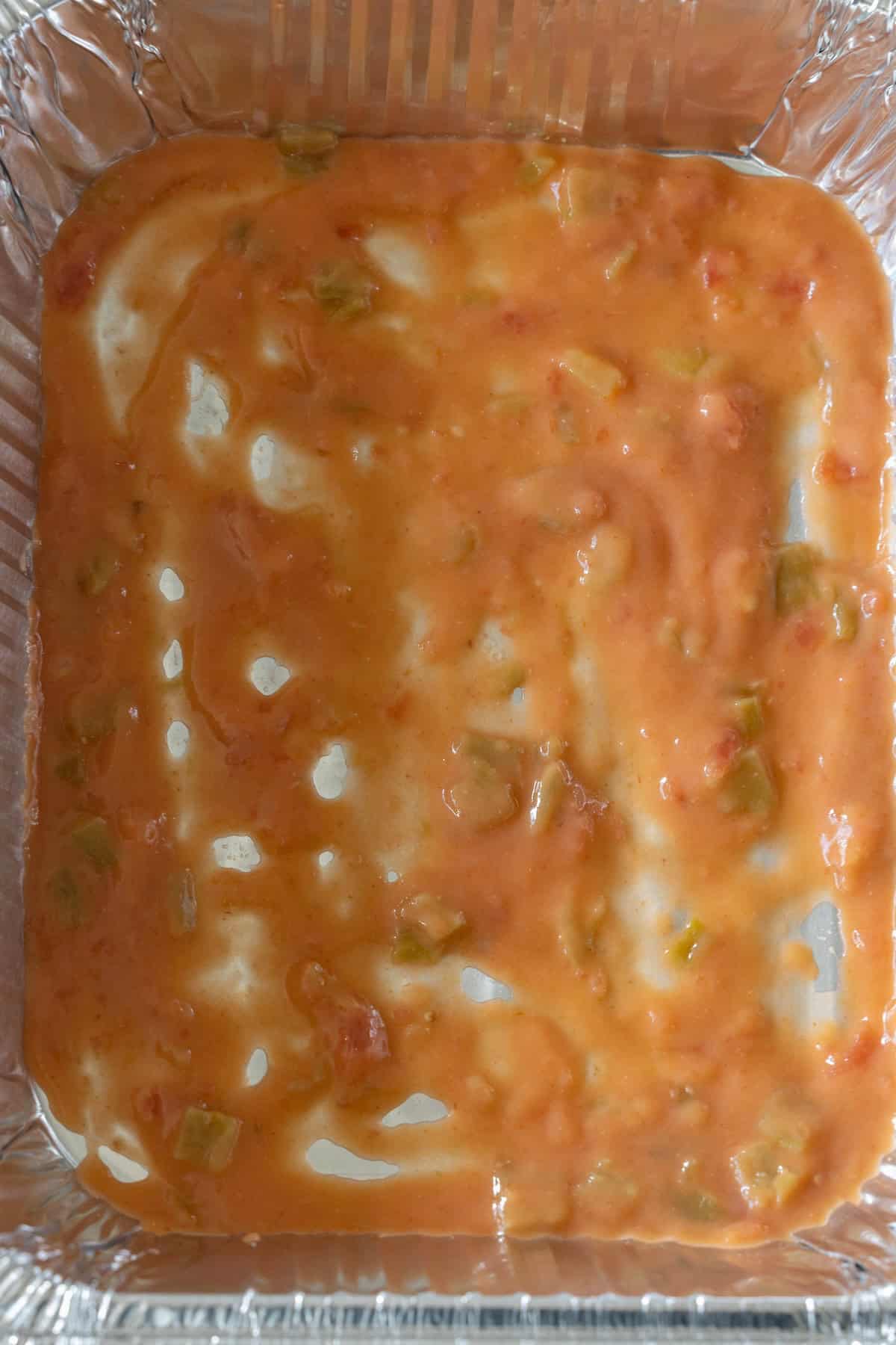 A close-up of a foil tray containing a creamy orange mixture with visible chunks of green and red vegetables—perfect for leftover turkey enchilada pie.