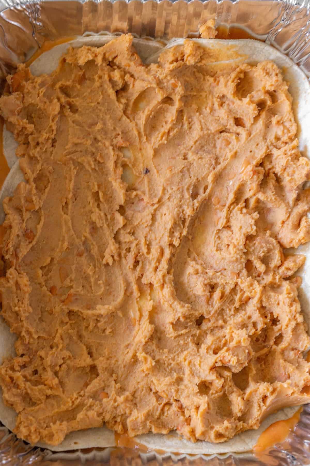 A layer of refried beans spread evenly over a tortilla base in a rectangular foil tray, perfect for building a delicious enchilada pie.