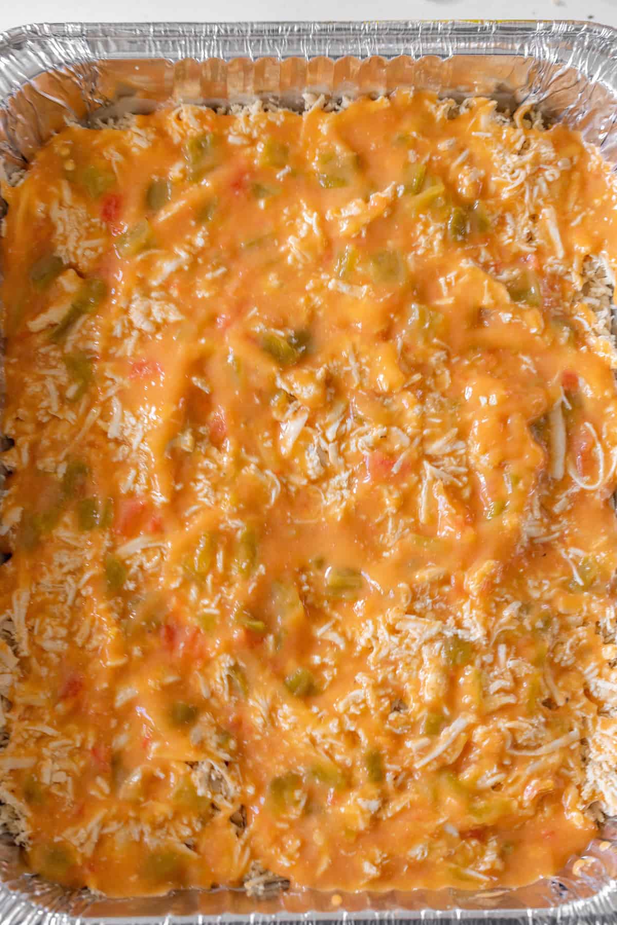 A close-up of a foil tray filled with an unbaked Turkey Enchilada casserole mixture, layered with shredded cheese, sauce, and diced vegetables.