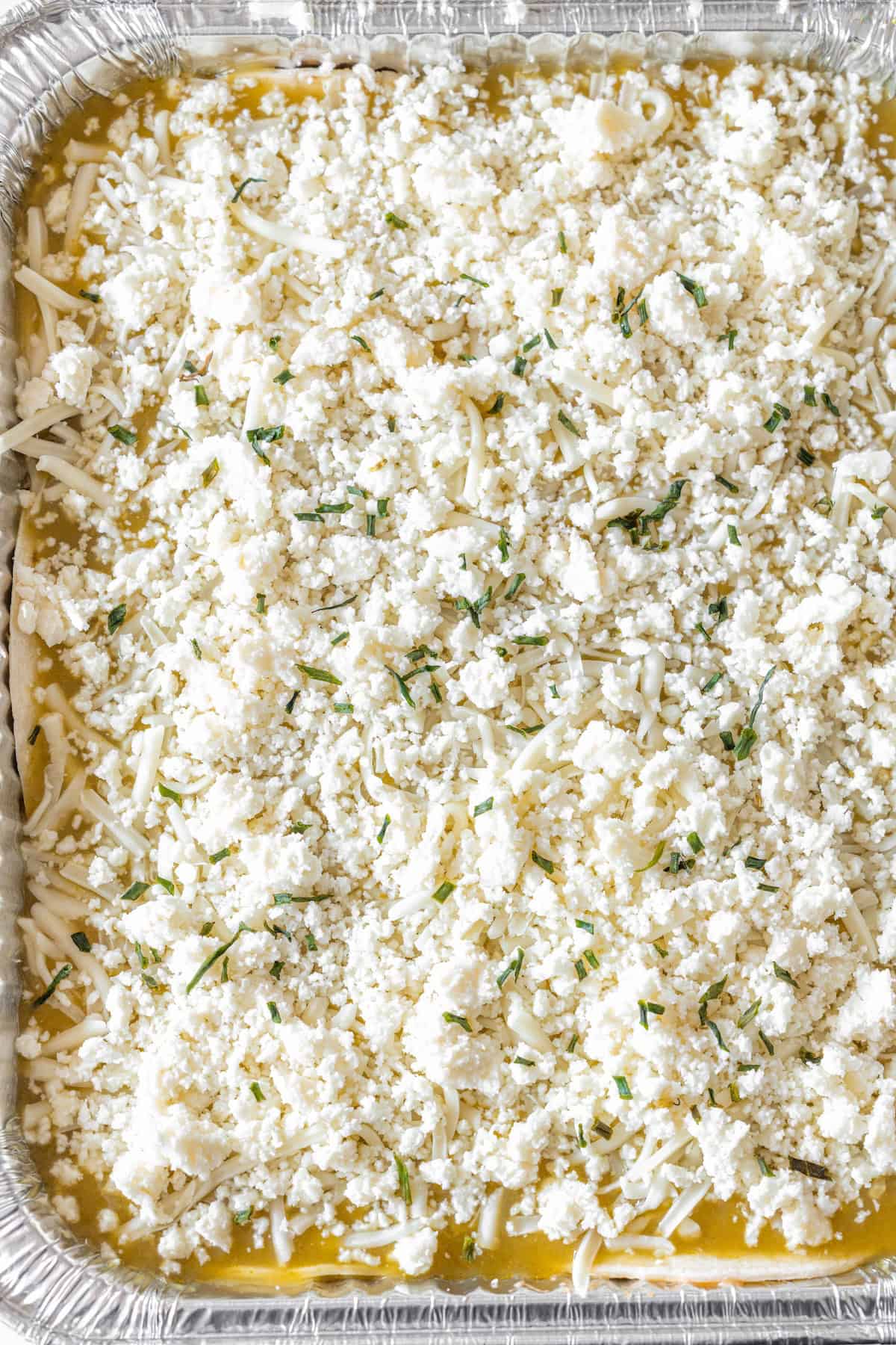 Rectangular foil tray filled with an unbaked enchilada pie, topped with crumbled cheese, shredded cheese, and herbs—perfect for using up leftover turkey.