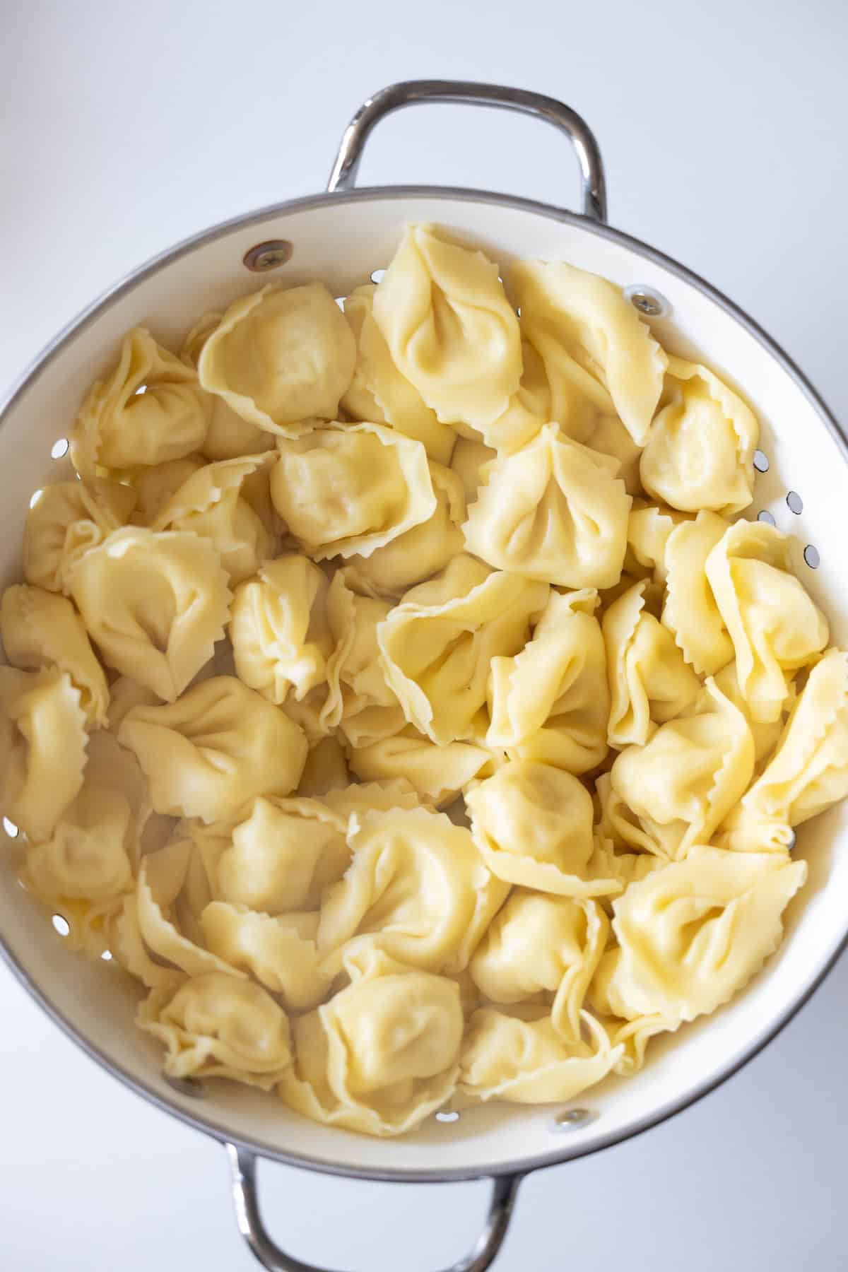 A metal colander filled with cooked cheese tortellini on a white background, perfect for your next Marry Me Tortellini recipe.
