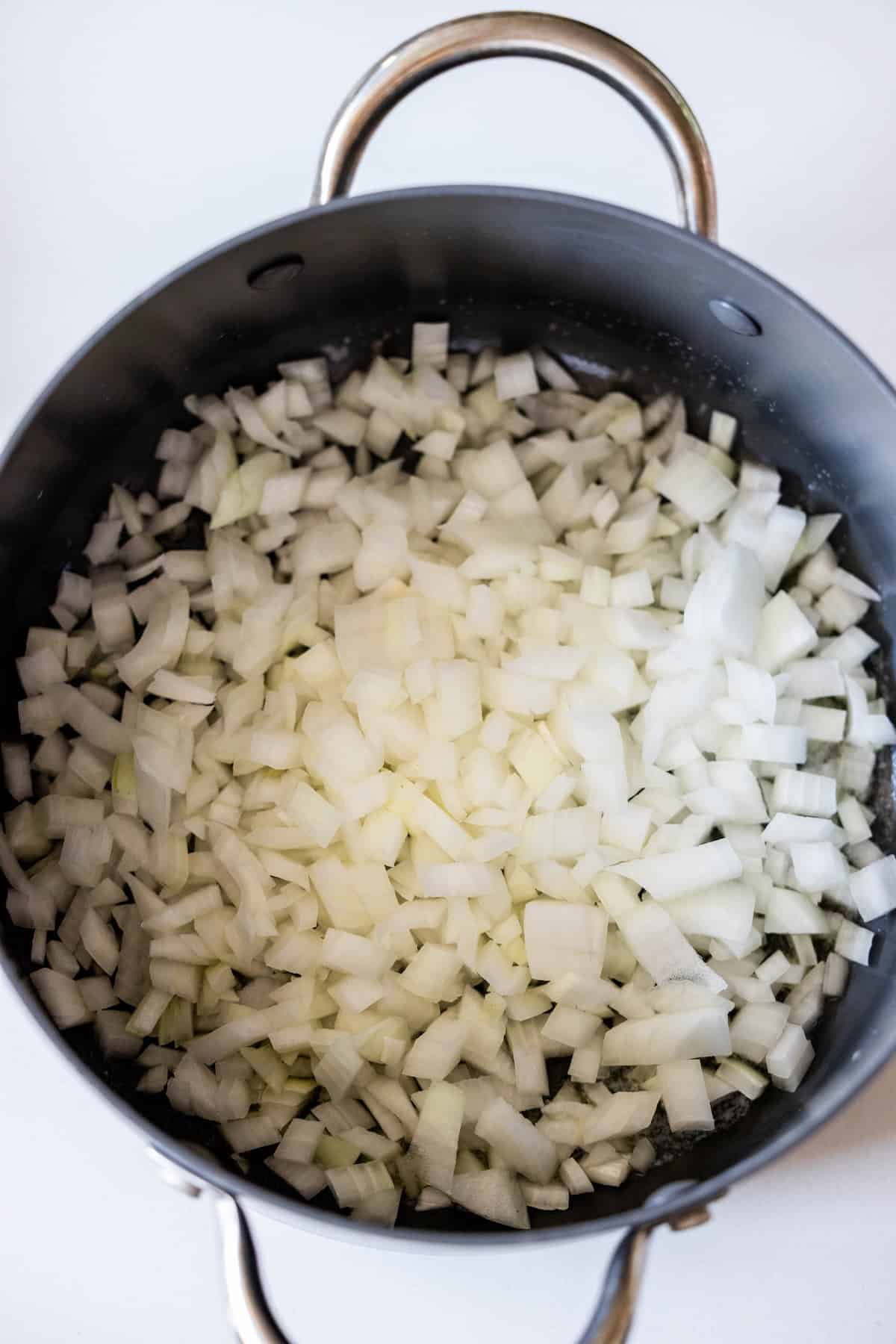 Chopped white onions in a black pot with metal handles, ready to be cooked for your favorite Marry Me Tortellini recipe.