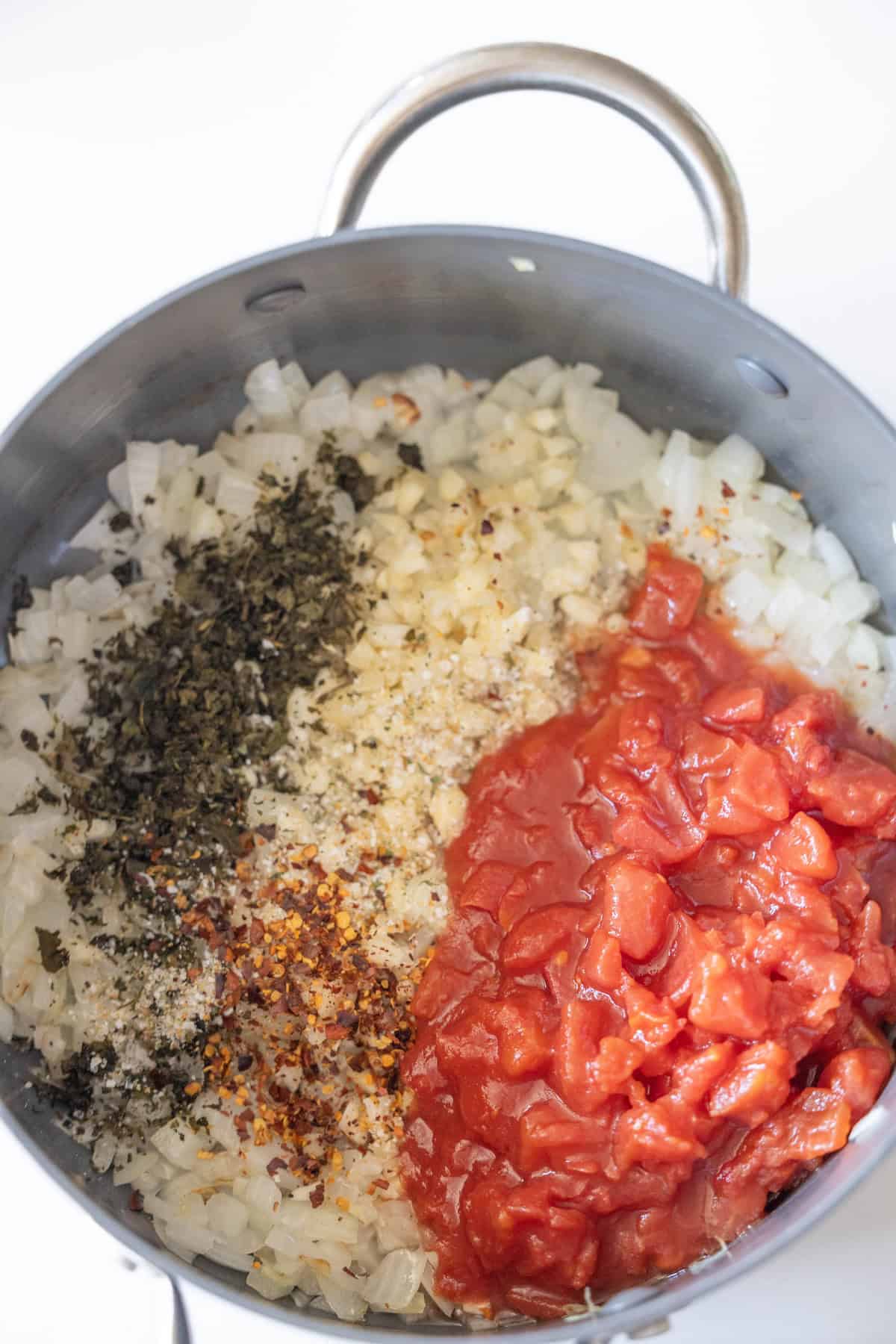 A pot with chopped onions, minced garlic, dried herbs, red pepper flakes, and diced tomatoes—all ready to be mixed for a flavorful Marry Me Tortellini recipe.