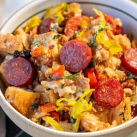 A bowl filled with bread pieces, sliced pepperoni, chopped peppers, and herbs, mixed together in a savory stuffing dish.