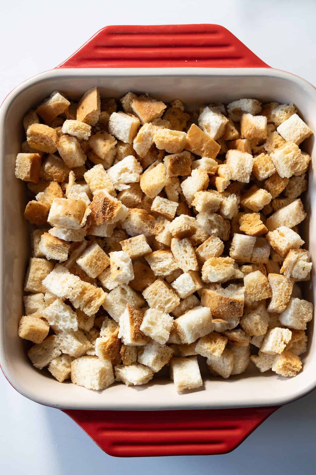 A baking dish filled with evenly cut bread cubes for stuffing, showing a mix of white and brown pieces, placed on a white surface.