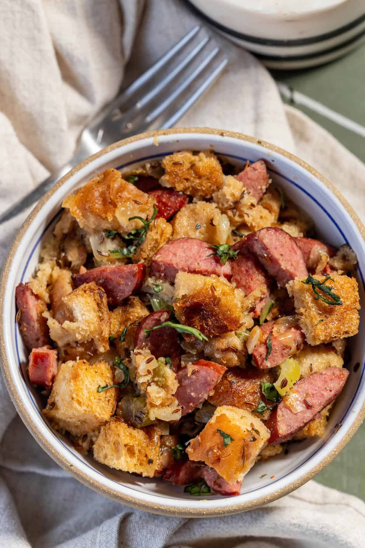 A bowl of bread stuffing mixed with Polish Kielbasa slices, herbs, and diced vegetables, placed on a light cloth with a fork beside it.