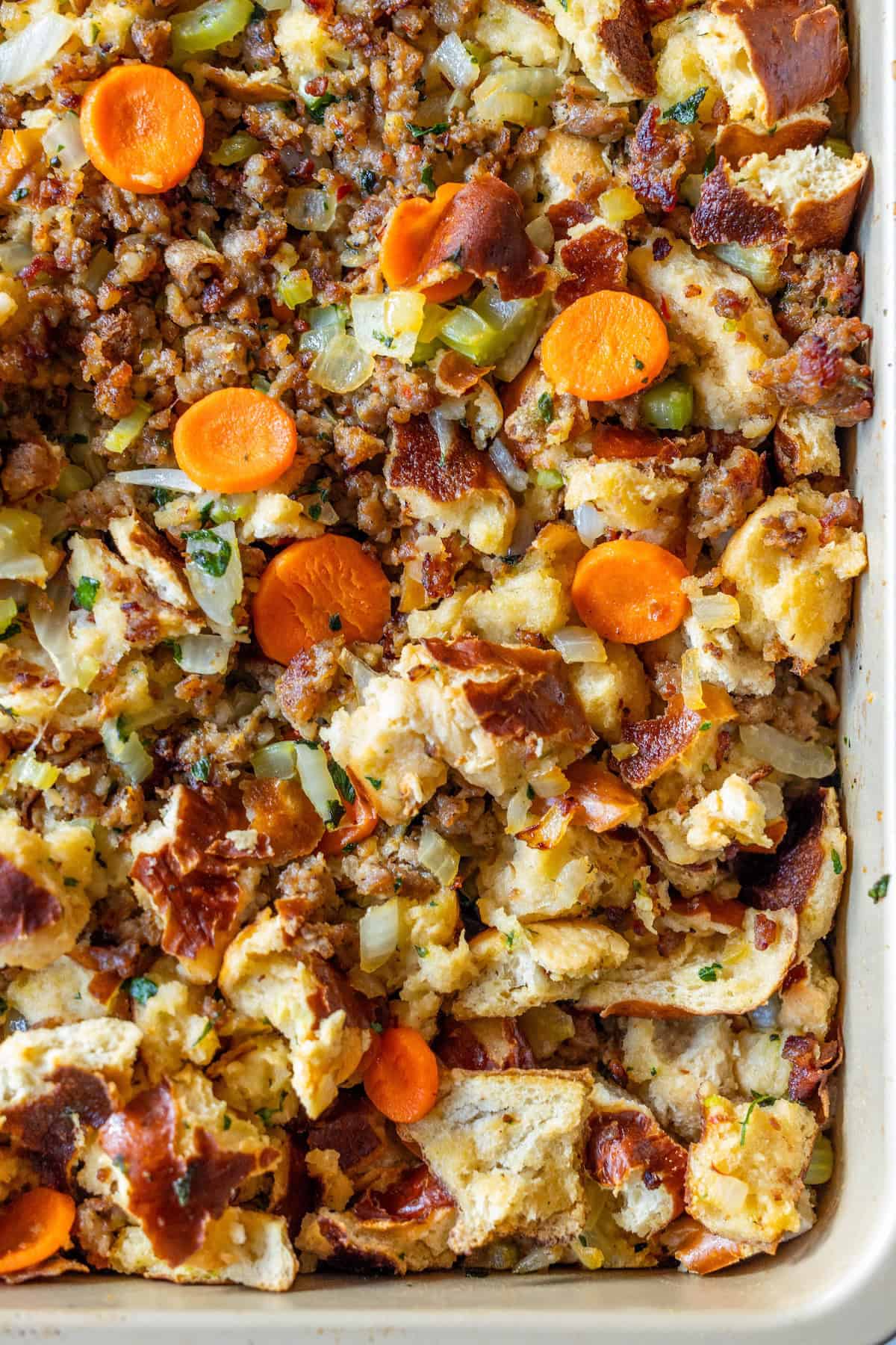 A close-up of a baked sausage stuffing dish with chunks of bread, sliced carrots, sausage, onions, celery, herbs, and toasted pretzel pieces.