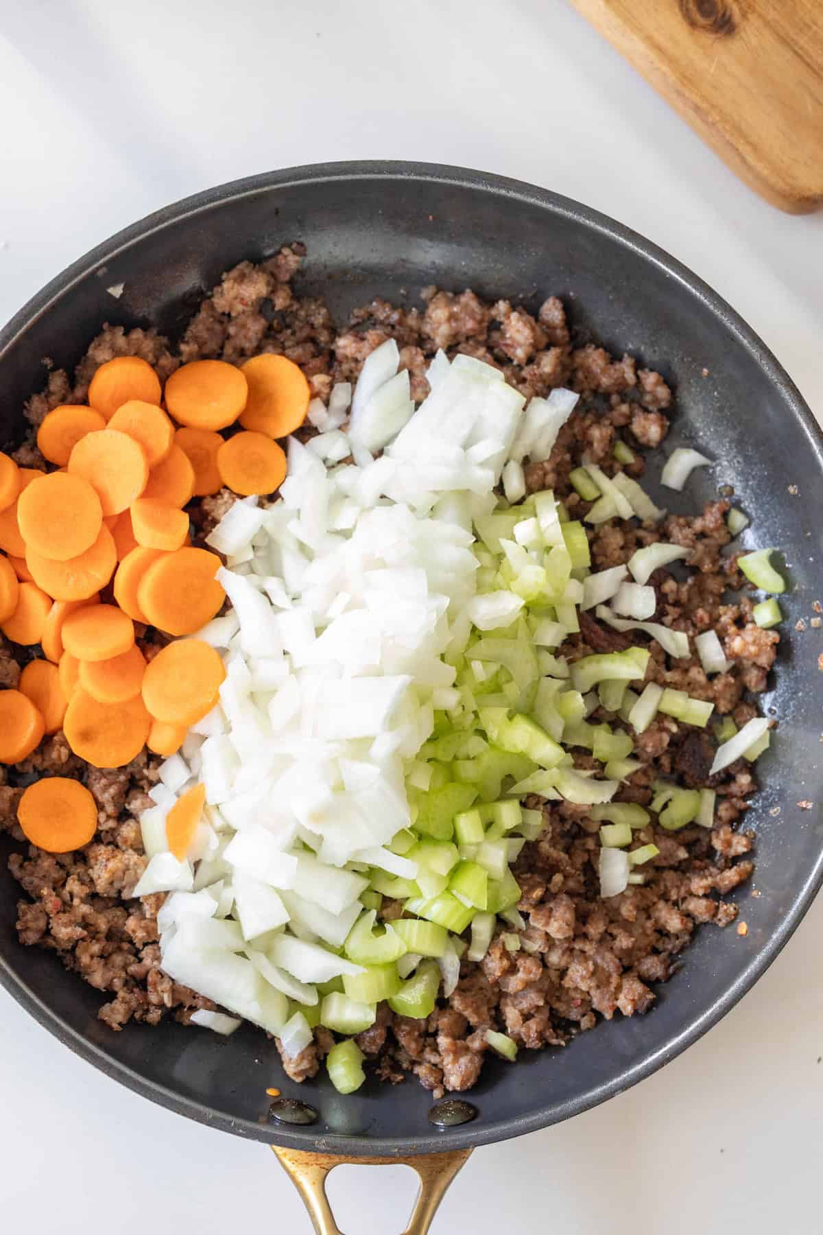 A frying pan with cooked ground meat, diced onions, chopped celery, and sliced carrots on a white surface—perfect as a base for pretzel stuffing.